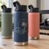 Three insulated stainless steel water bottles in navy, sage and coral, the navy bottle laser engraved with a silver palm tree and wave design, in a Pompano Beach engraving shop